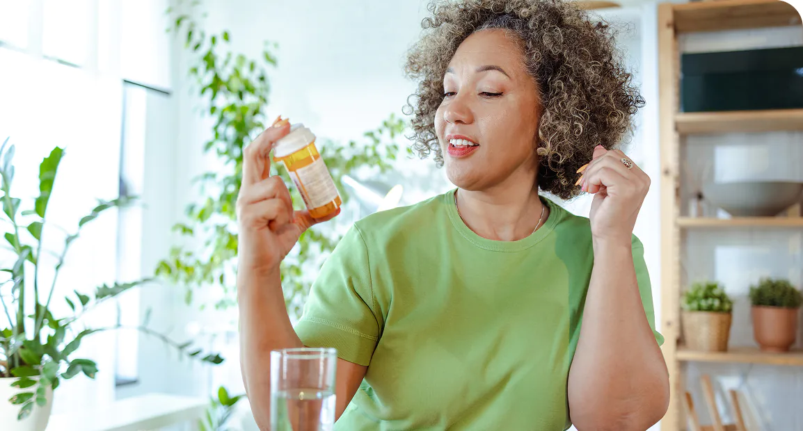 A woman in a green shirt holds a pill bottle and smiles, in a bright room with plants. A glass of water is on the table, evoking a health-focused mood.
