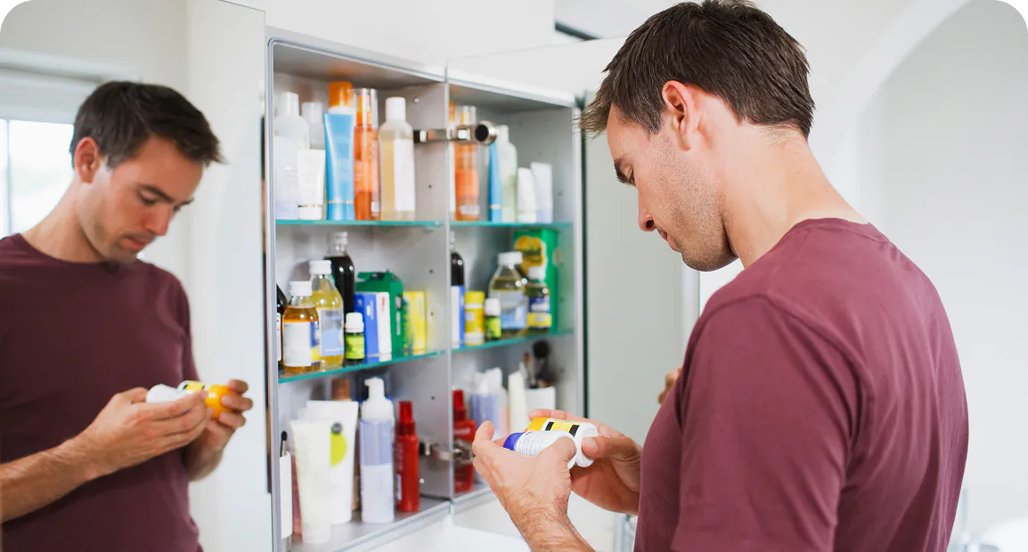 Man in a maroon shirt reads a label on a bottle in a bathroom, standing before a mirror cabinet filled with various toiletries.