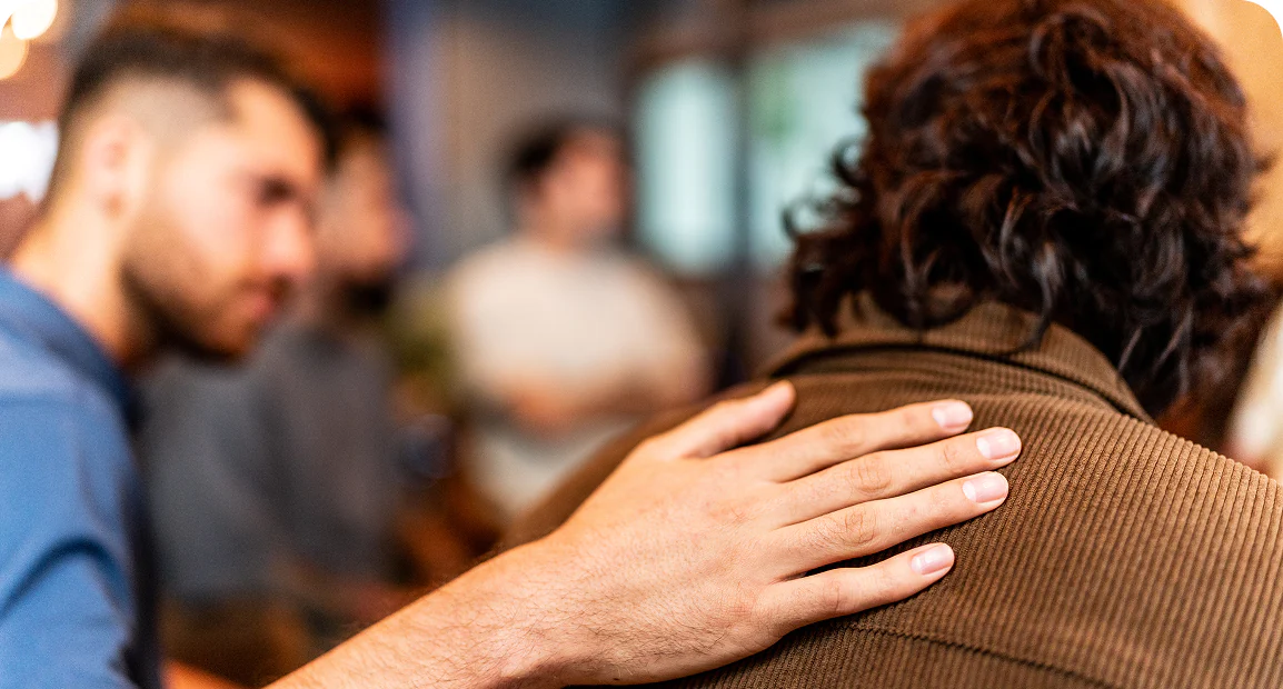 A close-up of a person placing a comforting hand on another's shoulder. The background is blurred, suggesting a supportive, emotional moment in a group setting.